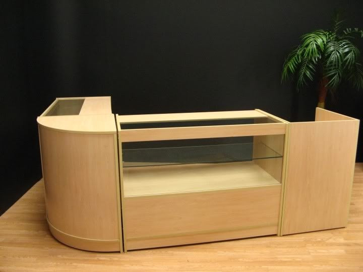 Bancone Cosmo in legno chiaro con finitura liscia, design curvo, ripiano in vetro e spazi di stoccaggio laterali, ideale per negozi