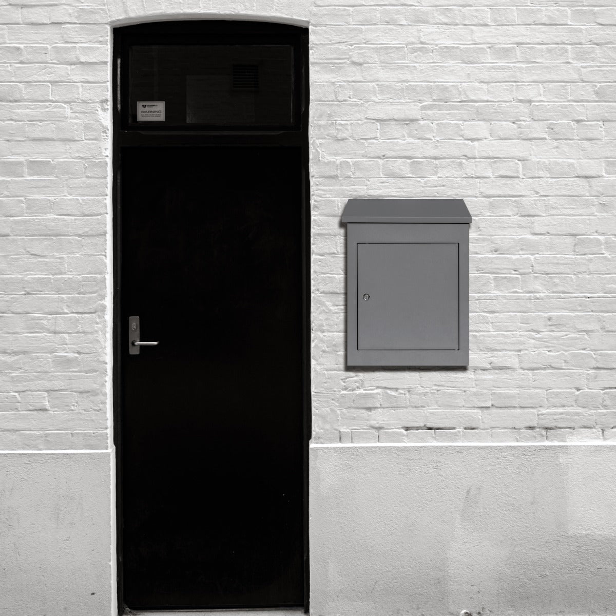 Cassetta postale grigia antifurto per pacchi, montata su muro bianco, con finitura liscia e design moderno, accanto a porta nera