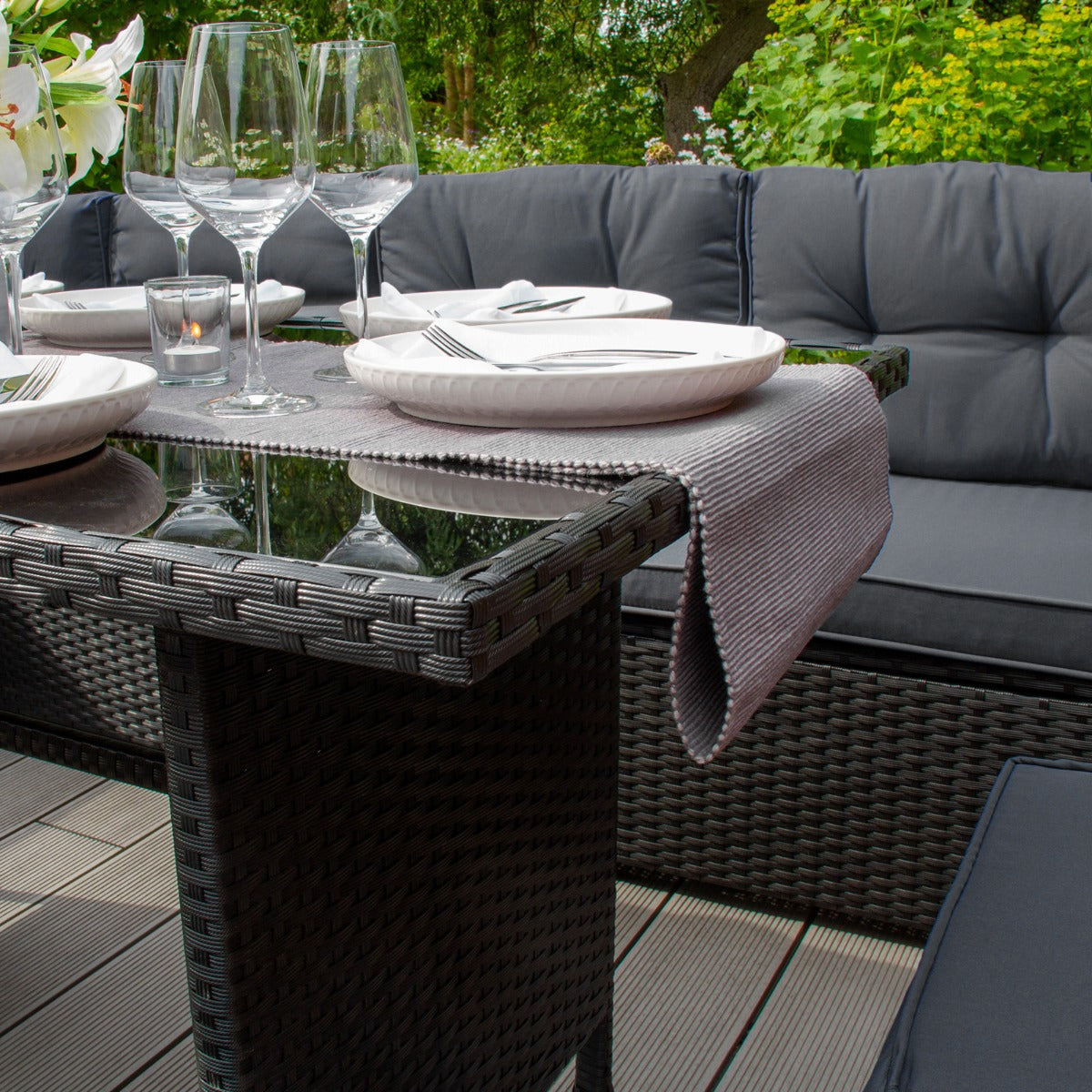Set ad angolo in rattan nero con cuscini grigi, tavolo in vetro, piatti bianchi e bicchieri eleganti su un patio verdeggiante