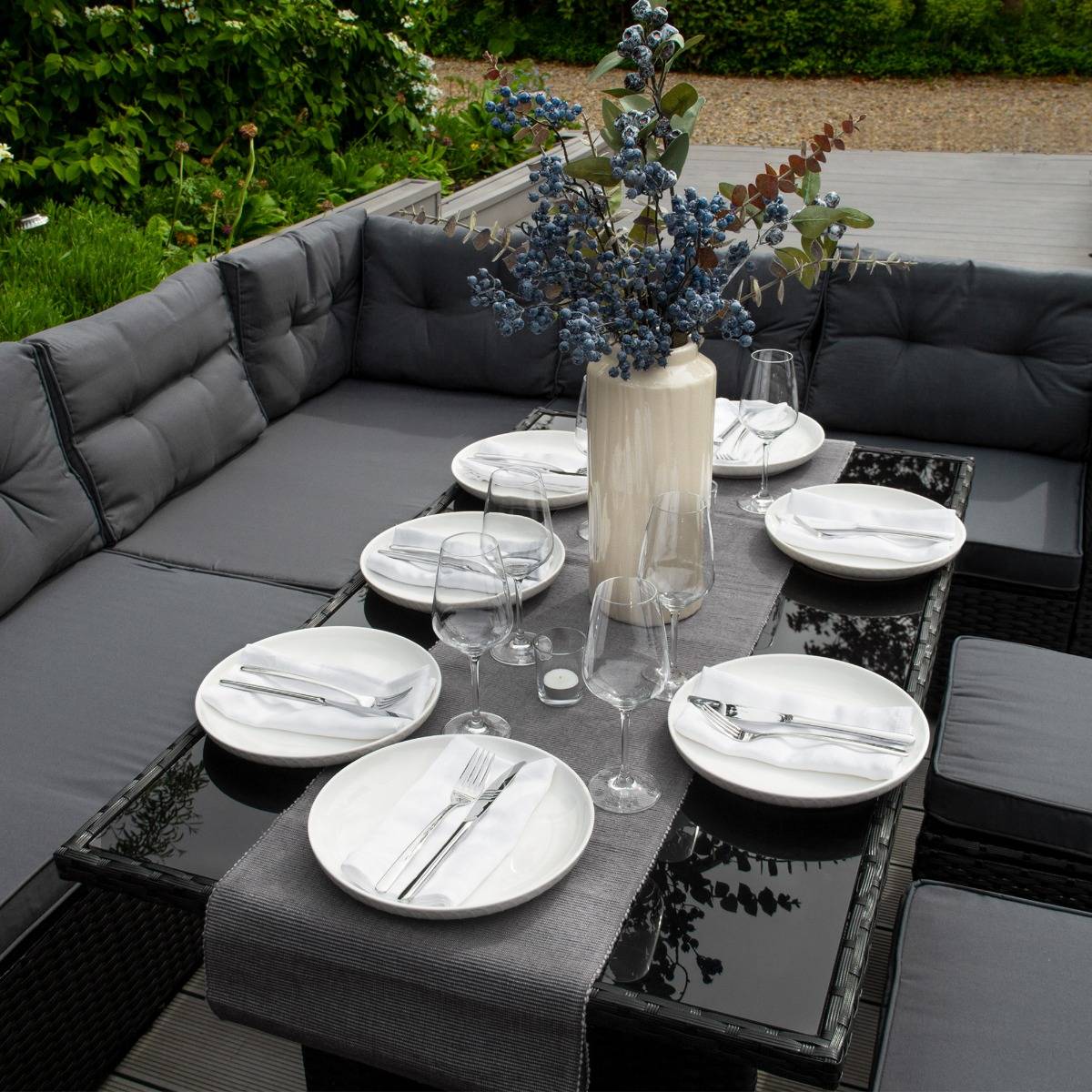 Set ad angolo in rattan nero con cuscini grigi, tavolo in vetro, piatti bianchi e un vaso con fiori blu al centro