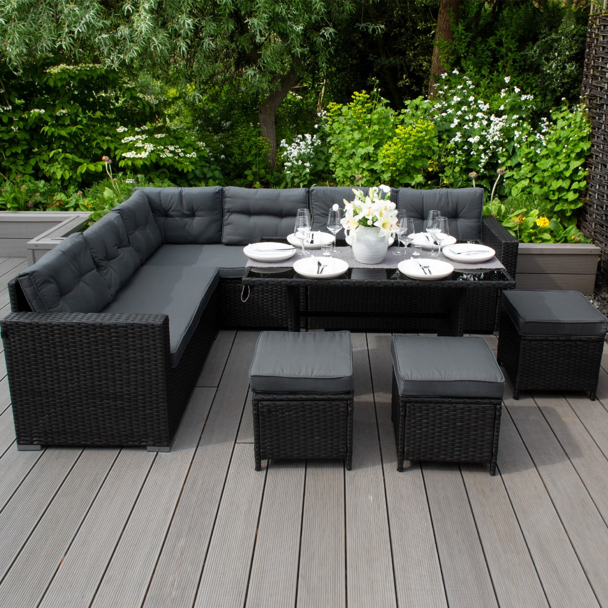 Set ad angolo in rattan nero con cuscini grigi, tavolo centrale e sgabelli, circondato da un giardino verde lussureggiante