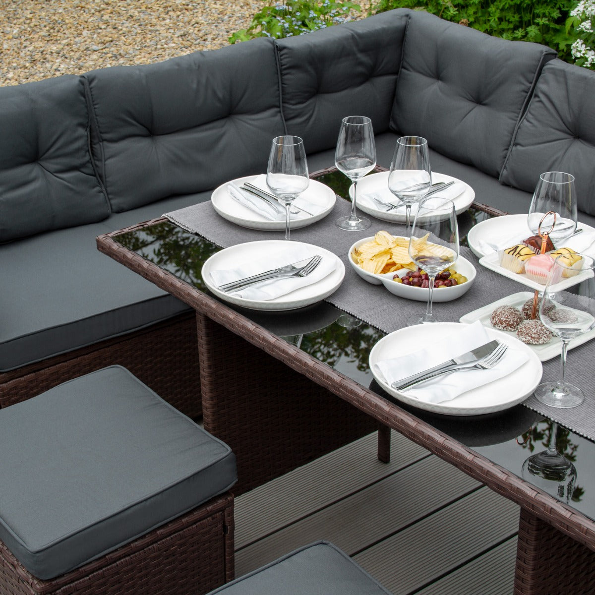 Set ad angolo in rattan marrone con cuscini grigi, tavolo in vetro, piatti bianchi e bicchieri, decorato con snack e frutta