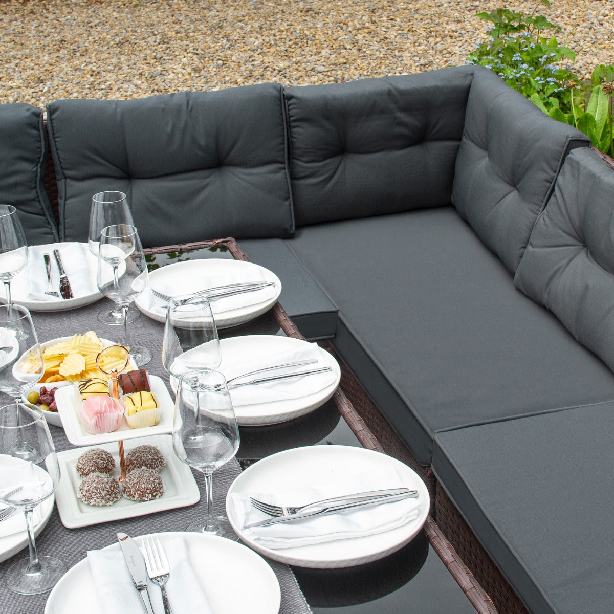 Set ad angolo in rattan marrone con cuscini grigi, tavolo apparecchiato con piatti bianchi e cibo, ambiente esterno elegante