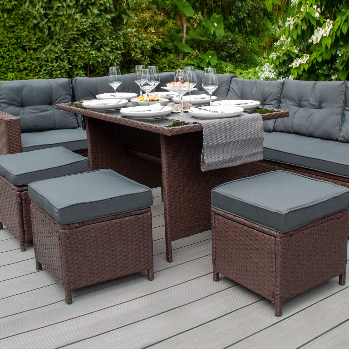 Set ad angolo in rattan marrone con cuscini grigi, tavolo centrale e stoviglie eleganti su un patio circondato da verde