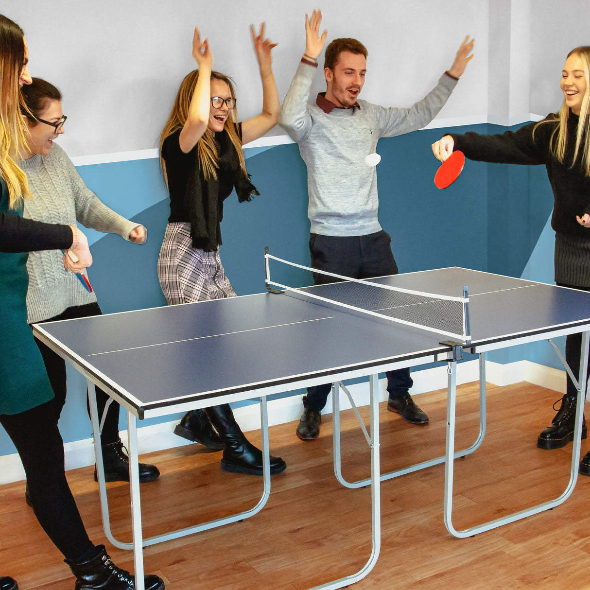 Tavolo da ping pong pieghevole blu con accessori, circondato da persone sorridenti che giocano e si divertono in un ambiente luminoso