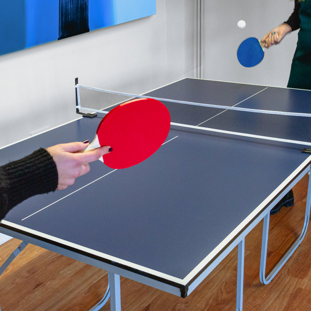 Tavolo da ping pong pieghevole blu con rete, racchette rosse e blu, in un ambiente luminoso con pareti chiare e arte moderna sullo sfondo