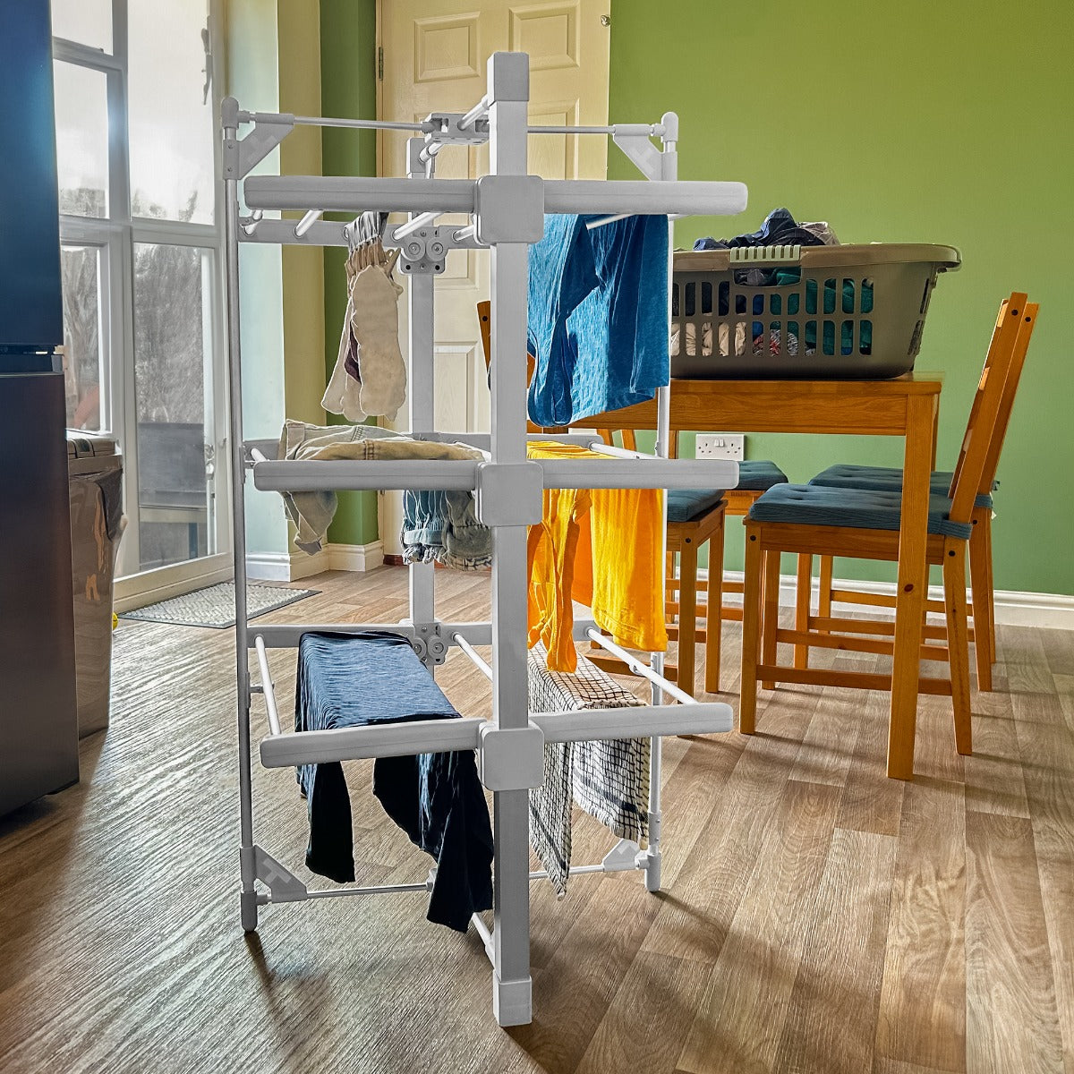 Stendibiancheria elettrico riscaldato bianco con panni stesi, posizionato su un pavimento in legno, vicino a un tavolo e una cesta di biancheria