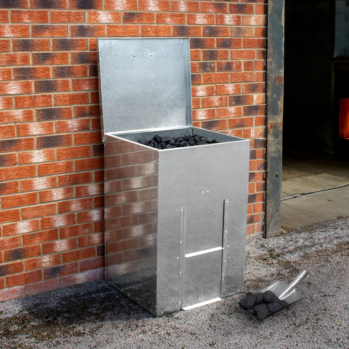 Bidone per carbone e legna in acciaio inox, con coperchio aperto, riempito di carbone, posizionato su una superficie di ghiaia vicino a un muro di mattoni rossi