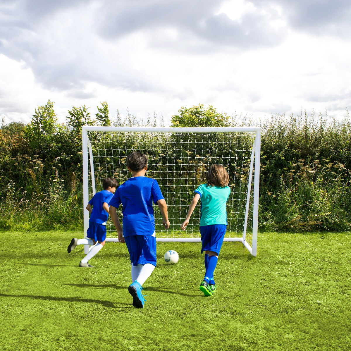 Tre bambini in maglietta blu e verde giocano a calcio davanti a una porta da 240cm x 180cm su un prato verde, cielo nuvoloso sullo sfondo