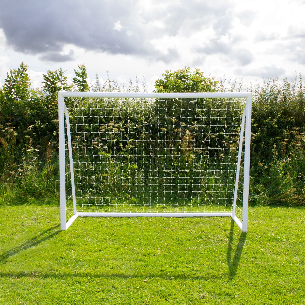 Porta da calcio bianca 240cm x 180cm con rete trasparente, posizionata su un prato verde, circondata da alberi e cielo nuvoloso