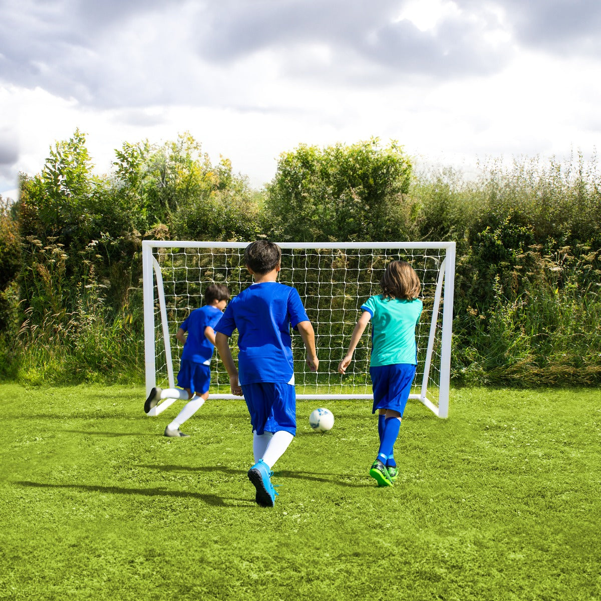 Tre bambini in maglietta blu e pantaloni corti corrono verso una porta da calcio bianca su un prato verde, con nuvole nel cielo