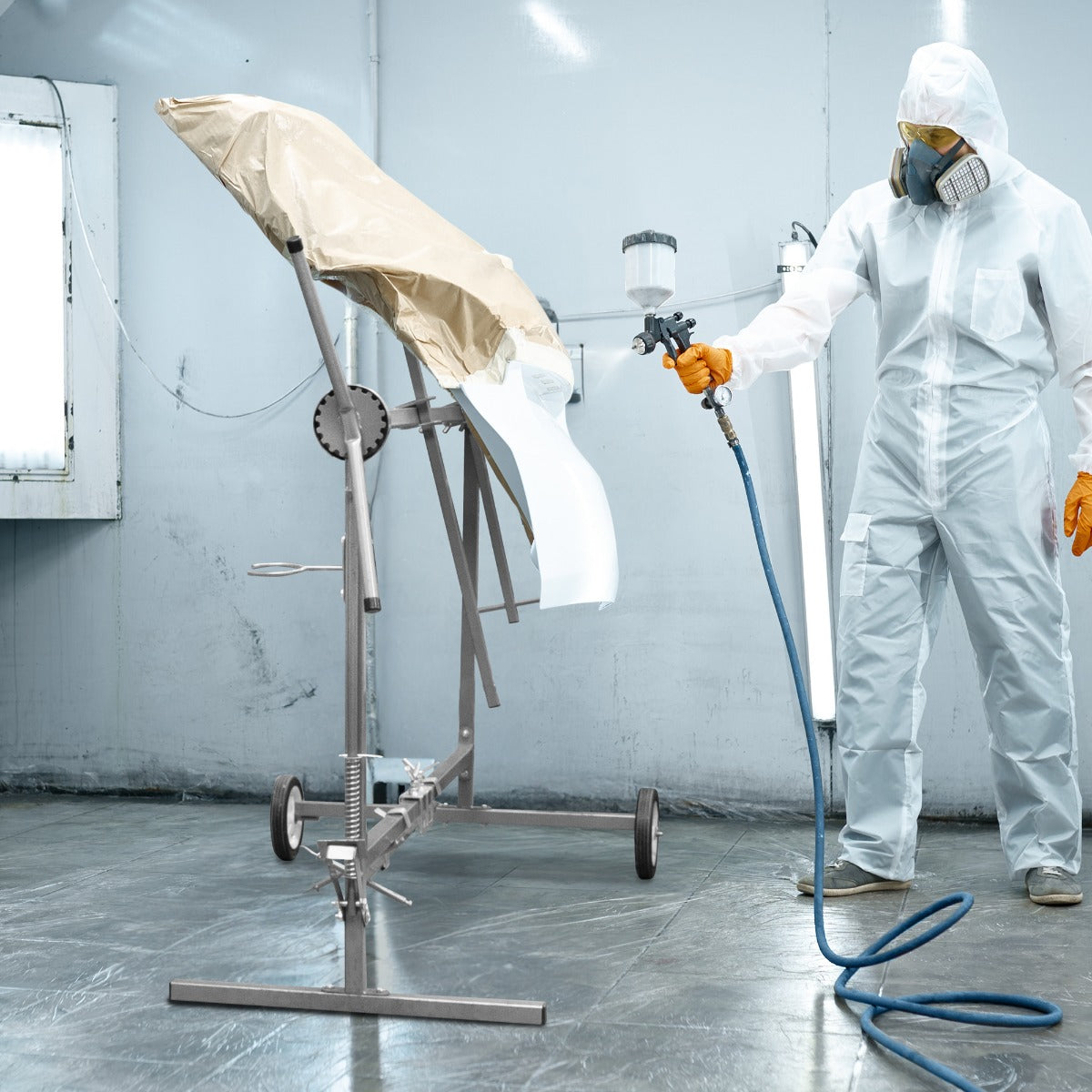 Supporto rotante per verniciatura in metallo grigio con ruote, utilizzato da un operatore in tuta bianca e maschera, mentre spruzza vernice