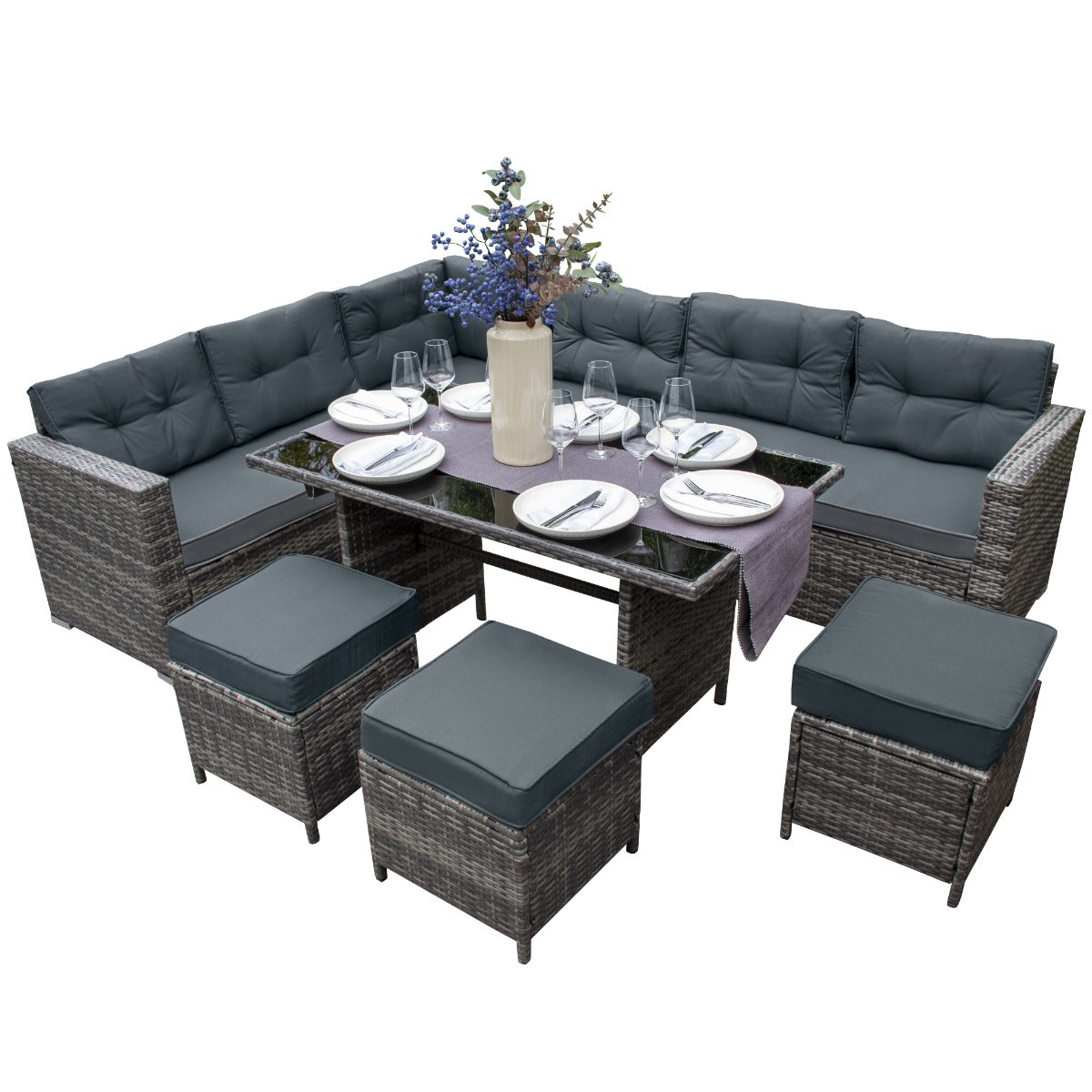 Set ad angolo in rattan grigio con cuscini morbidi, tavolo in vetro, decorato con fiori e piatti eleganti per un pranzo all'aperto