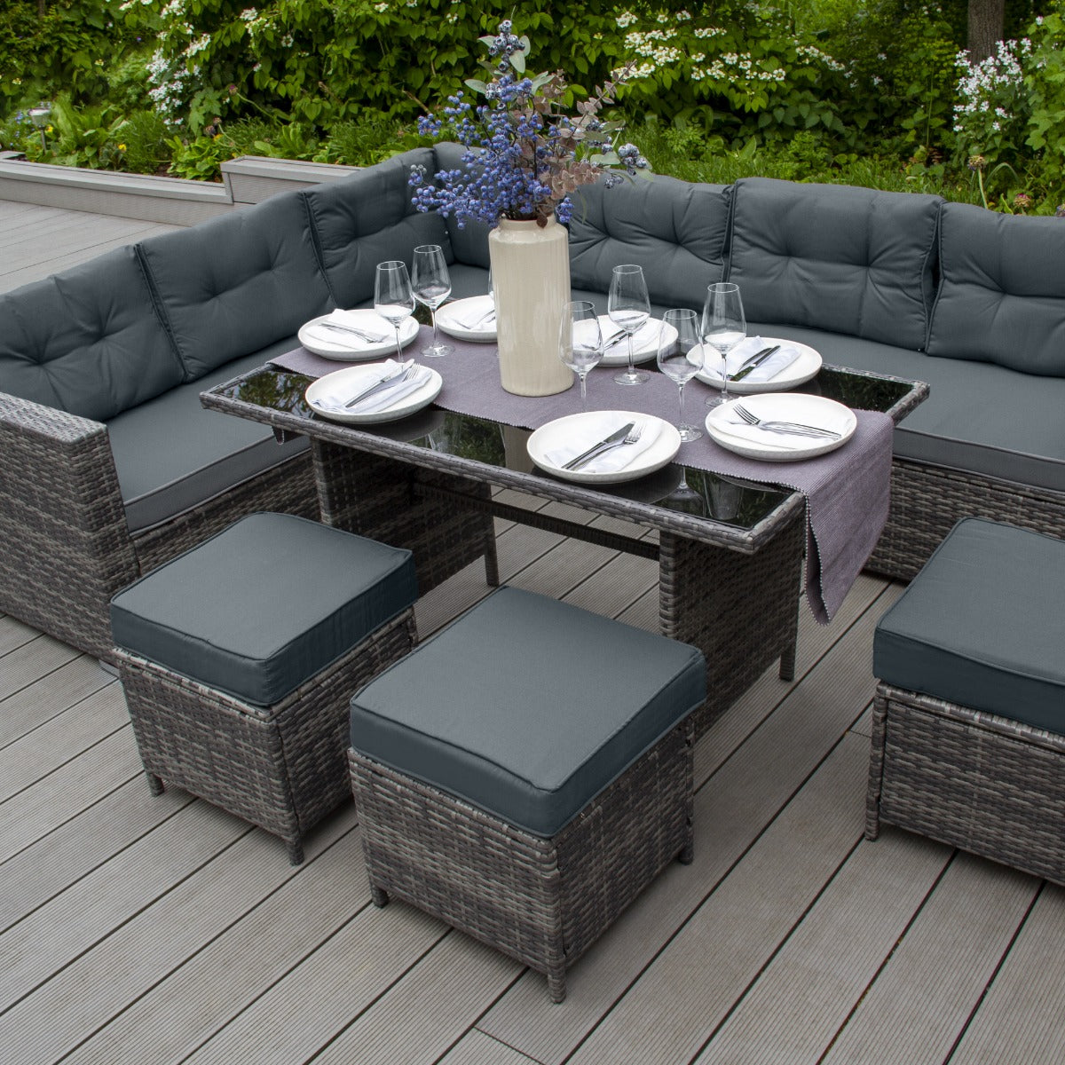 Set ad angolo in rattan grigio con cuscini scuri, tavolo in vetro e decorazione floreale, ideale per spazi esterni eleganti