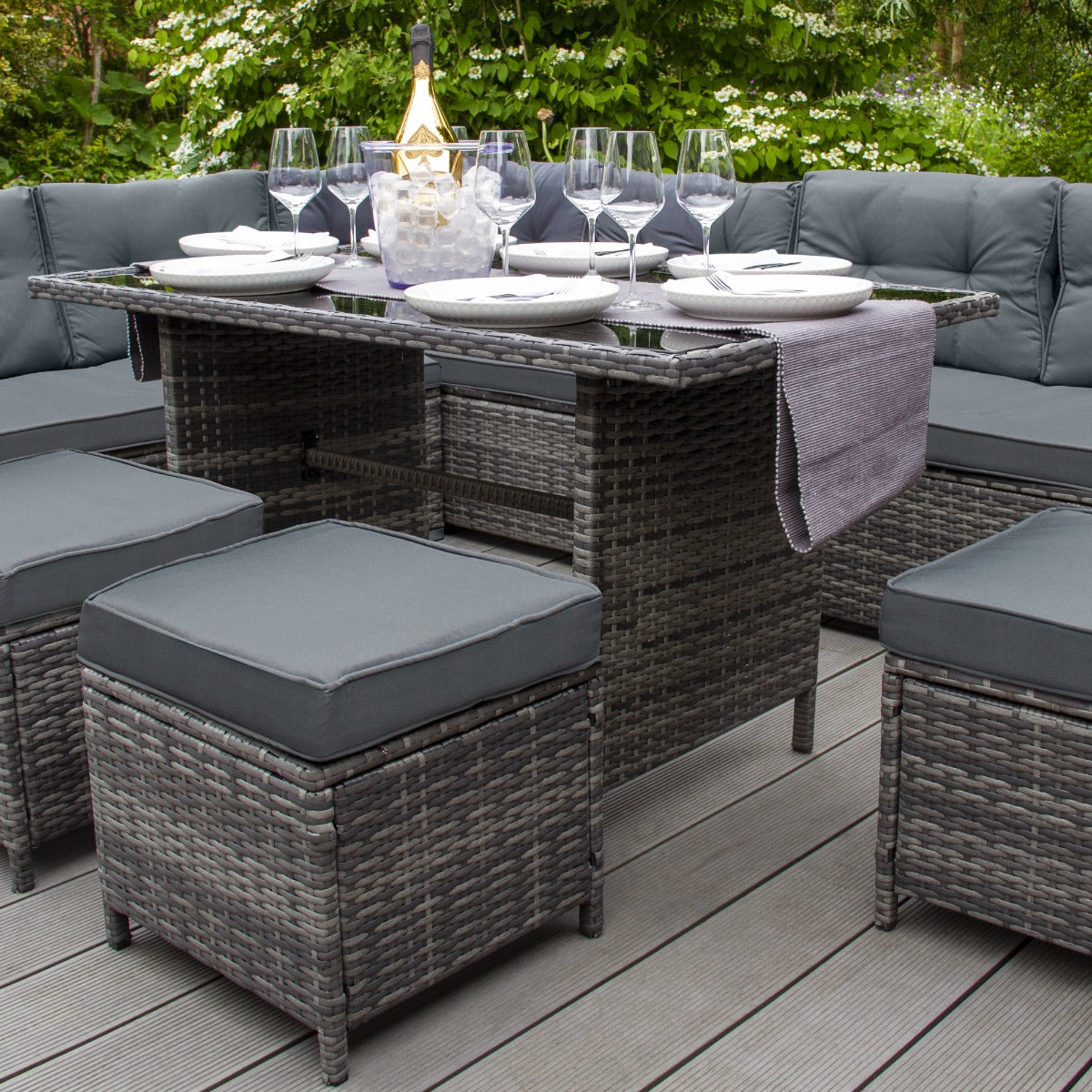 Set ad angolo in rattan grigio con cuscini scuri, tavolo centrale elegante e bicchieri da vino su un patio immerso nel verde
