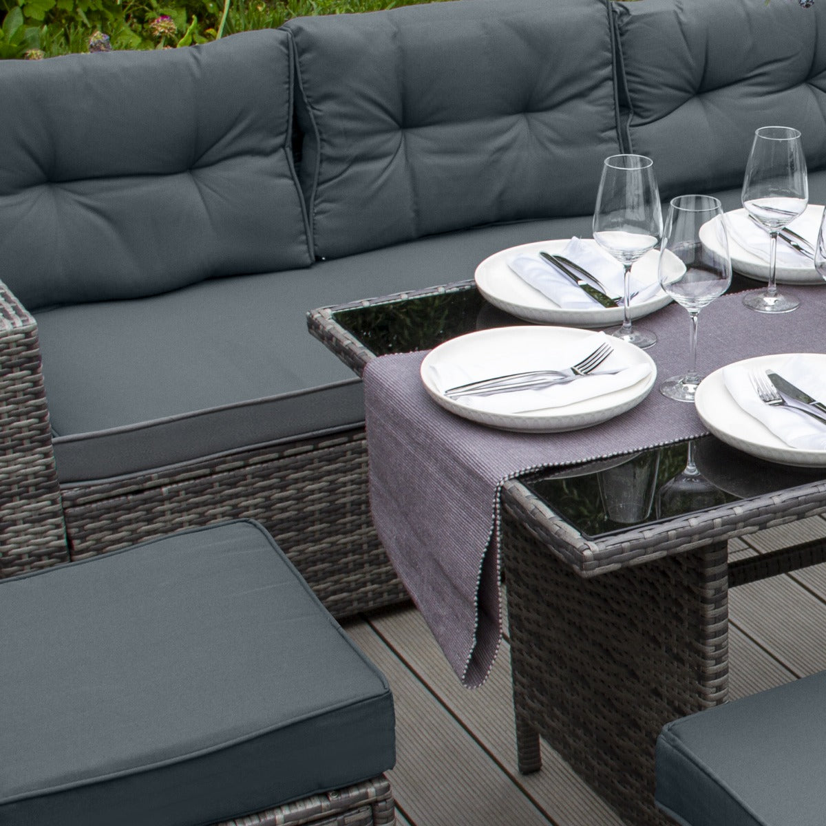 Set ad angolo in rattan grigio con cuscini morbidi, tavolo in vetro, piatti bianchi e bicchieri eleganti su un tavolo apparecchiato