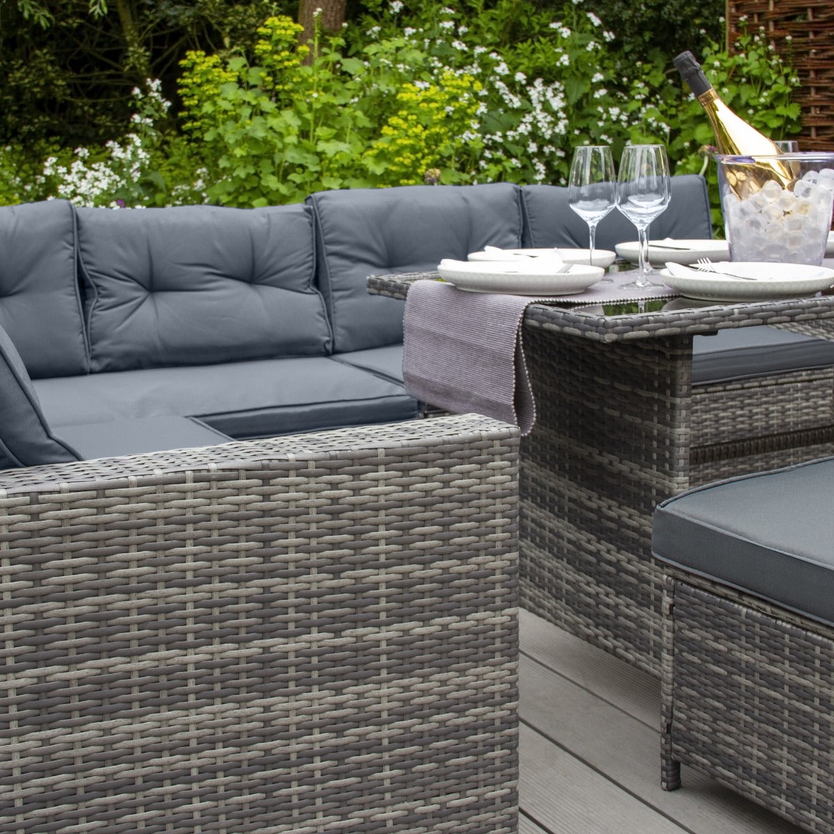 Set ad angolo in rattan grigio con cuscini imbottiti, tavolo elegante, bicchieri e bottiglia di champagne in un giardino verdeggiante