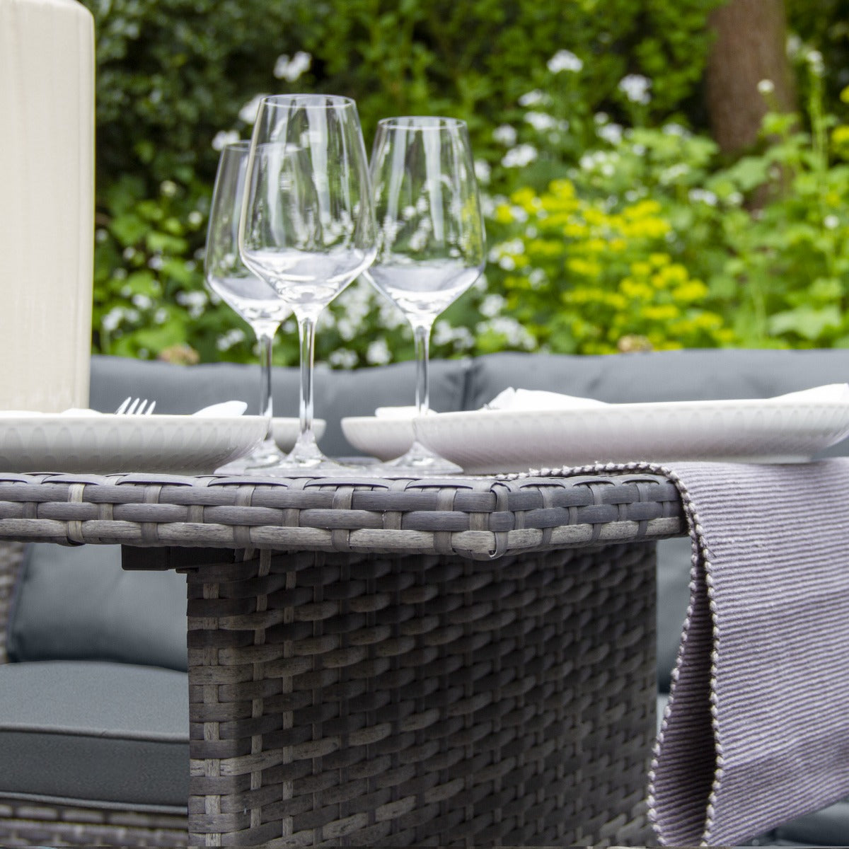 Set ad angolo in rattan grigio con tavolo, bicchieri di vetro e piatti, circondato da un giardino fiorito verde e luminoso