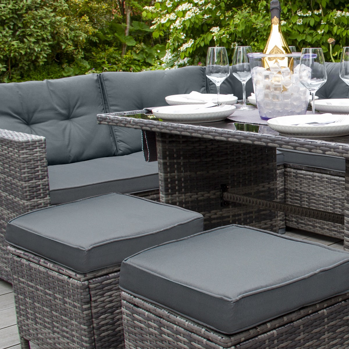 Set ad angolo in rattan grigio con cuscini coordinati, tavolo elegante e bicchieri di vetro, immerso in un ambiente verde e rigoglioso