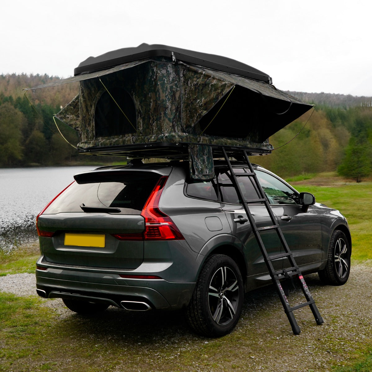 Tenda da tetto mimetica montata su un'auto grigia, con scala, immersa in un paesaggio naturale verde e sereno