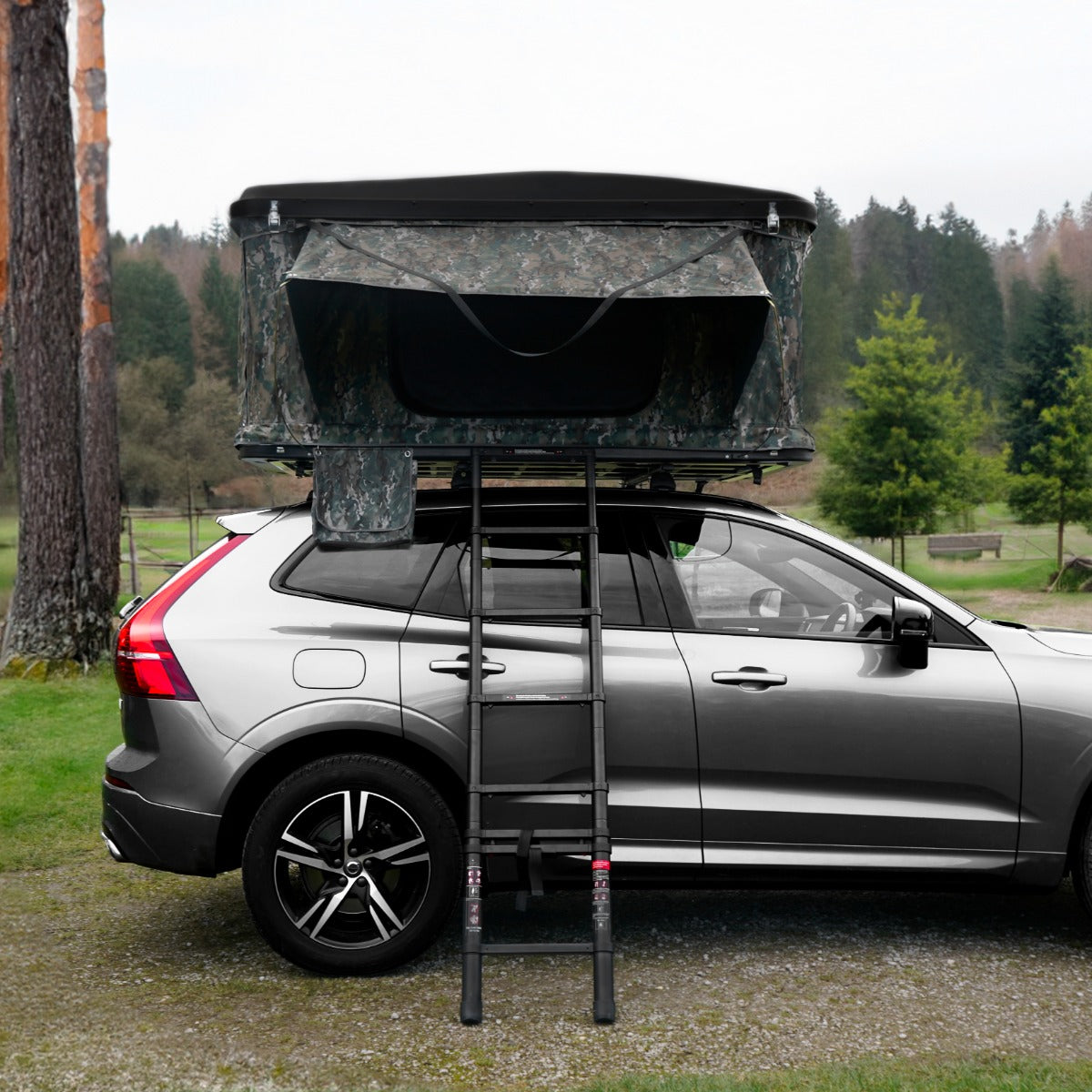 Tenda da tetto mimetica montata su un'auto grigia, con scala nera, circondata da alberi e un paesaggio naturale verde