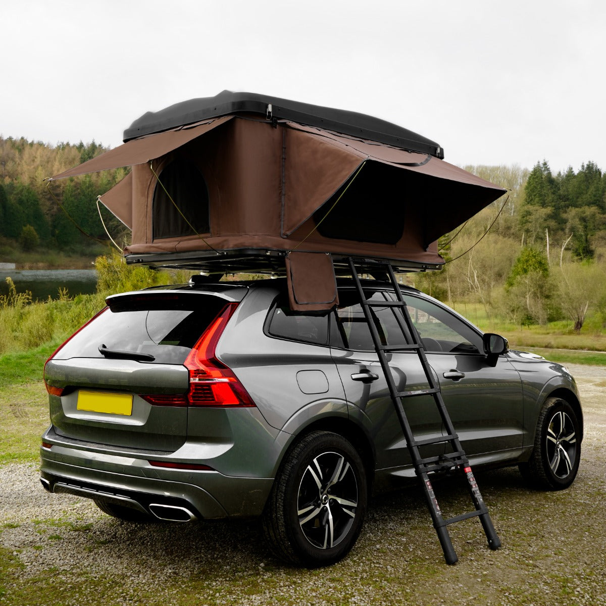 Tenda da tetto marrone montata su un'auto grigia, con aperture laterali e scala per l'accesso, immersa nella natura verde