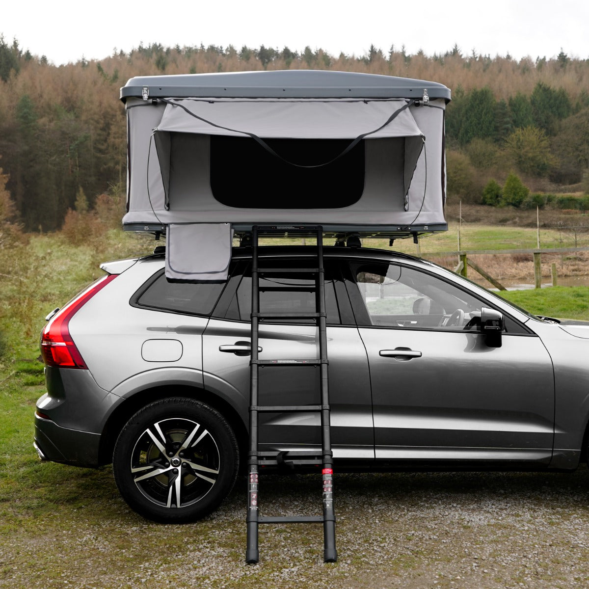 Tenda da tetto grigia montata su un'auto, con scala nera, in un paesaggio naturale verde e alberato sullo sfondo