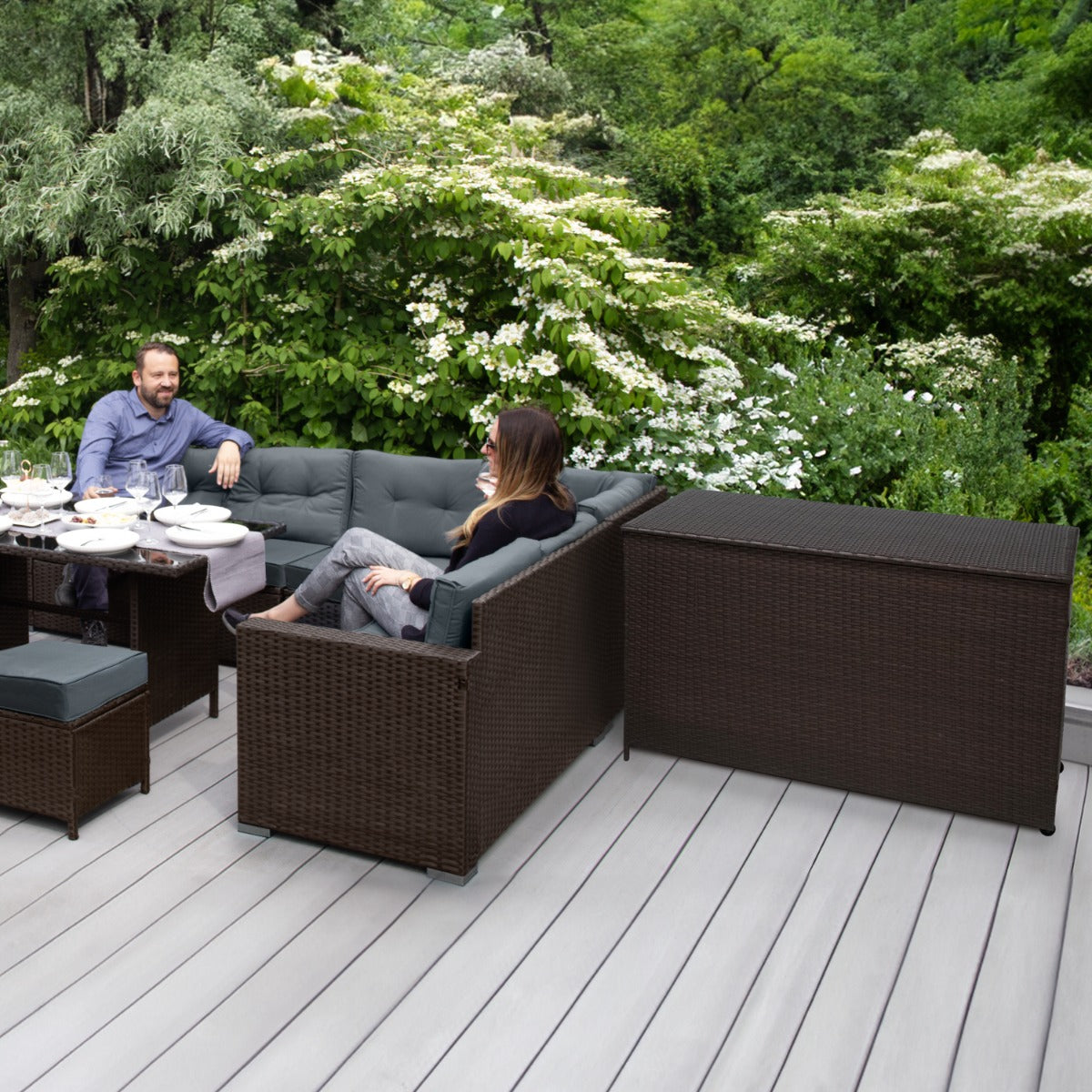 Set d'angolo in rattan marrone con cuscini grigi, tavolino e box per riporre i cuscini, immerso in un giardino verde lussureggiante