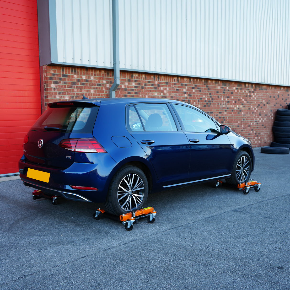 Pattini di posizionamento idraulici arancioni sotto una Volkswagen Golf blu, su una superficie grigia, con sfondo di mattoni rossi