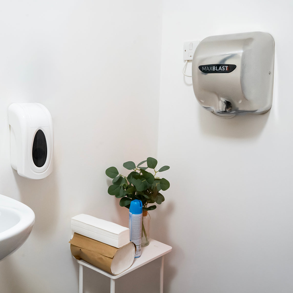 Asciugamani elettrico Maxblast grigio montato a parete, dispenser di sapone bianco e pianta verde su tavolino in un bagno moderno