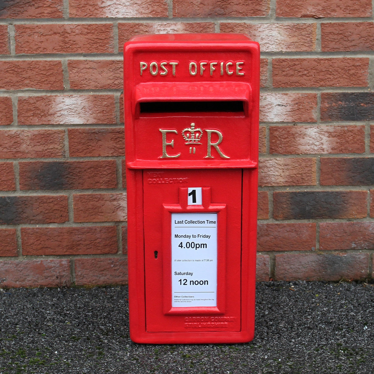 Cassetta postale britannica rossa con piedistallo, decorata con il monogramma ER e un avviso di raccolta su sfondo mattonato