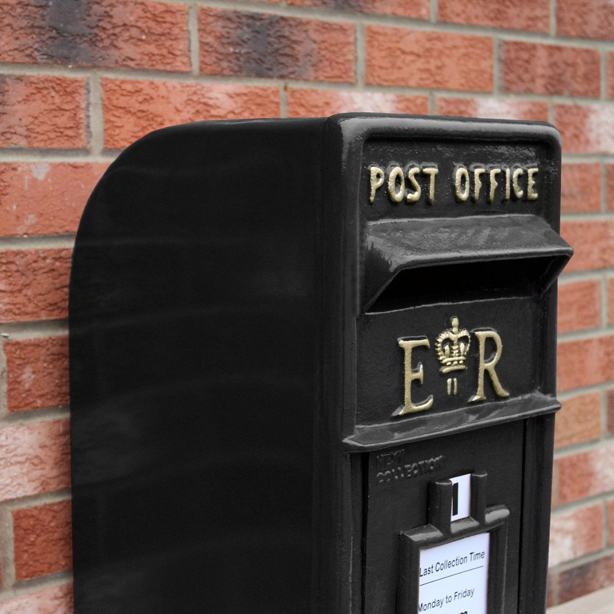Cassetta postale britannica nera con piedistallo, decorata con dettagli dorati e il simbolo ER, su sfondo di mattoni rossi
