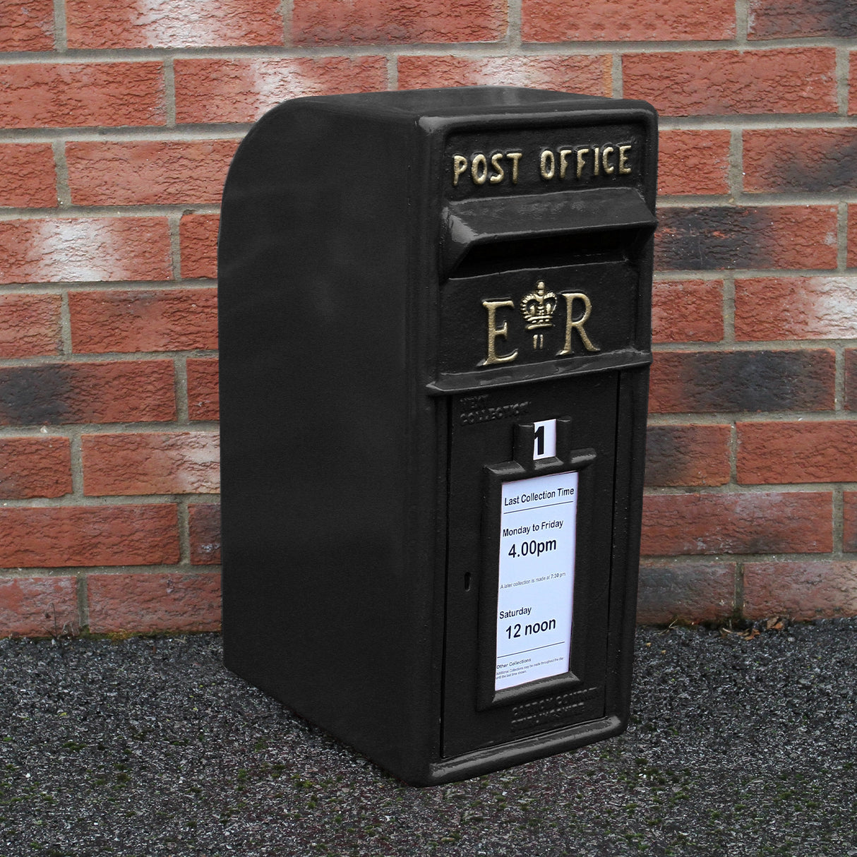 Cassetta postale britannica nera con piedistallo, decorata con dettagli dorati e scritta "POST OFFICE" su sfondo di mattoni rossi