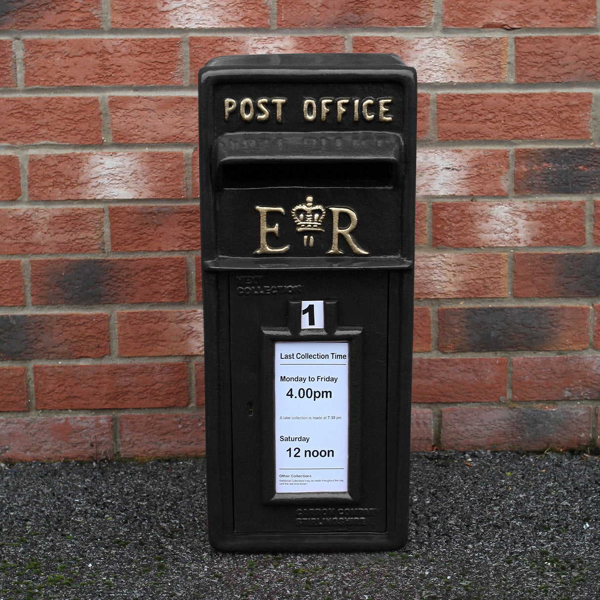 Cassetta postale britannica nera con piedistallo, dettagli dorati, iscrizioni chiare e avviso di raccolta affisso sulla parte anteriore