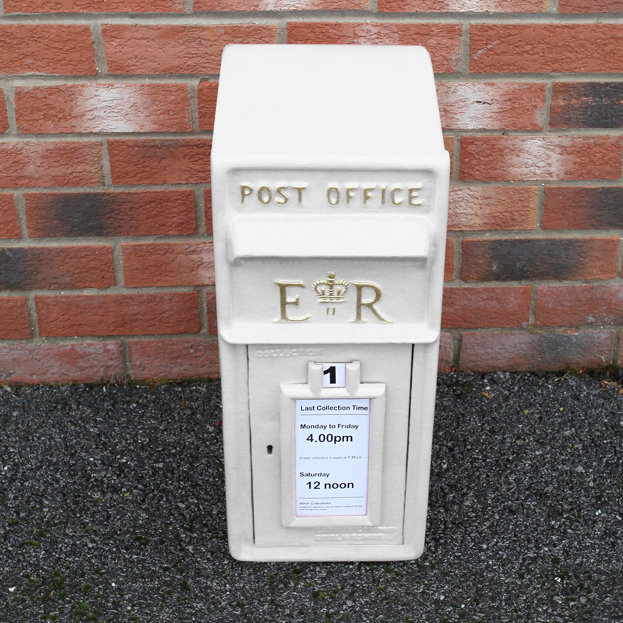Cassetta postale britannica bianca con dettagli dorati, scritta "POST OFFICE" e orari di raccolta su un muro di mattoni rossi