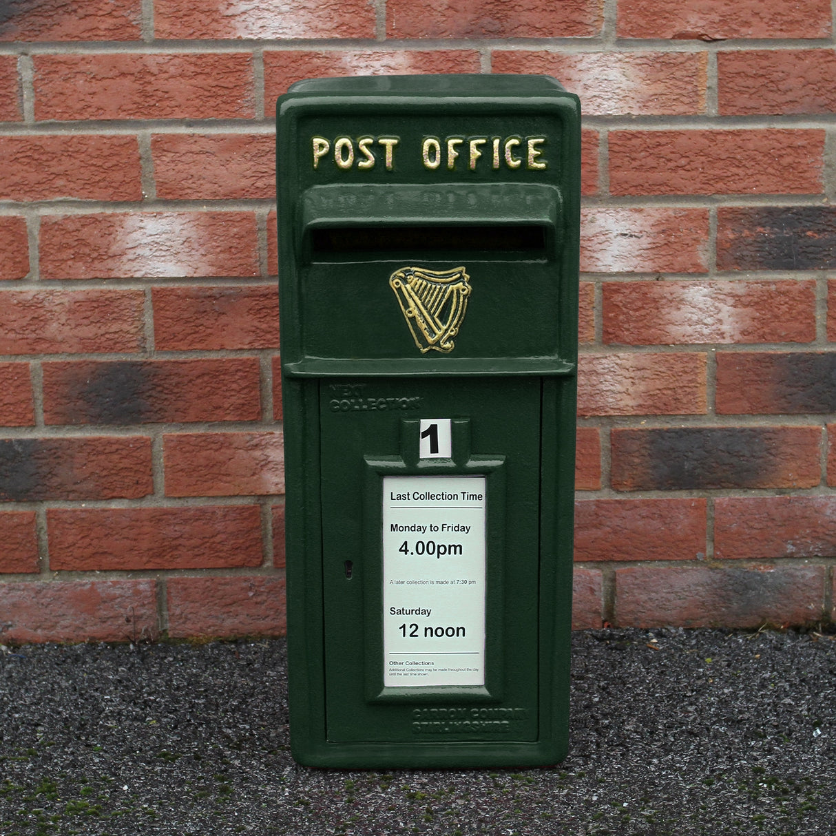 Cassetta postale britannica verde con dettagli dorati, logo arpa e avviso di raccolta su sfondo di mattoni rossi