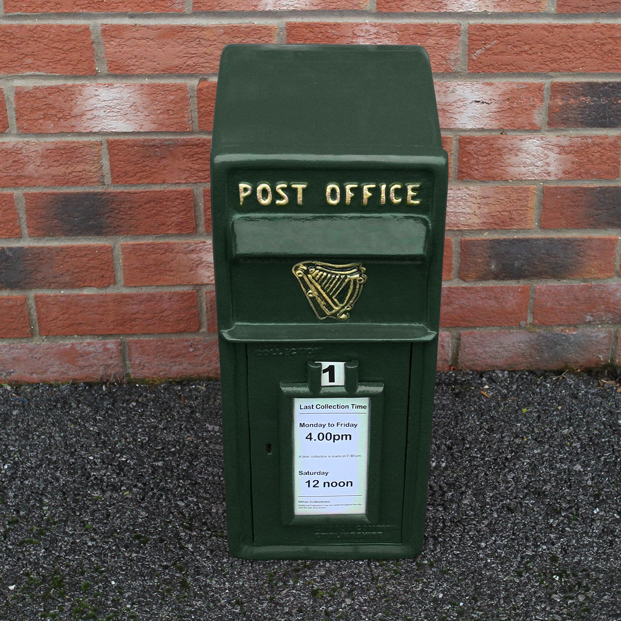 Cassetta postale britannica verde con dettagli dorati, logo arpa, orari di raccolta e base nera su sfondo di mattoni rossi