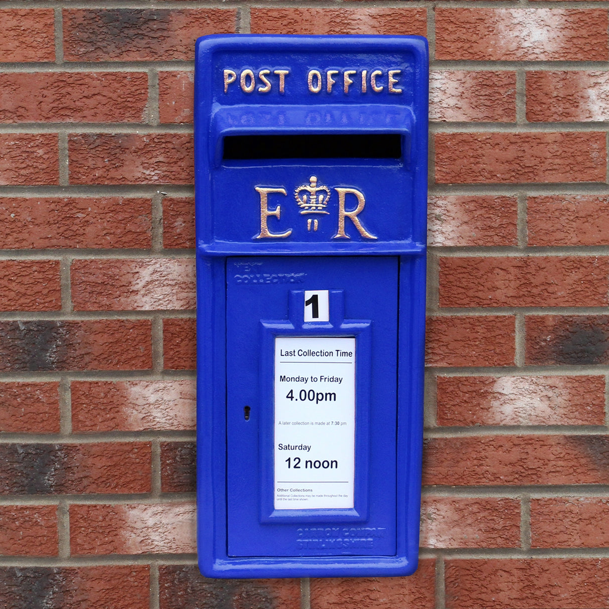Cassetta postale britannica blu con dettagli dorati, logo ER e orari di raccolta su un muro di mattoni rossi