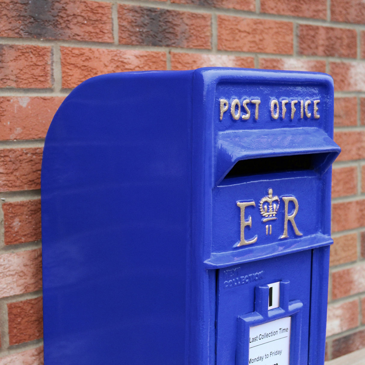 Cassetta postale britannica blu brillante con dettagli dorati, logo ER e fessura per la posta, appoggiata su un muro di mattoni rossi