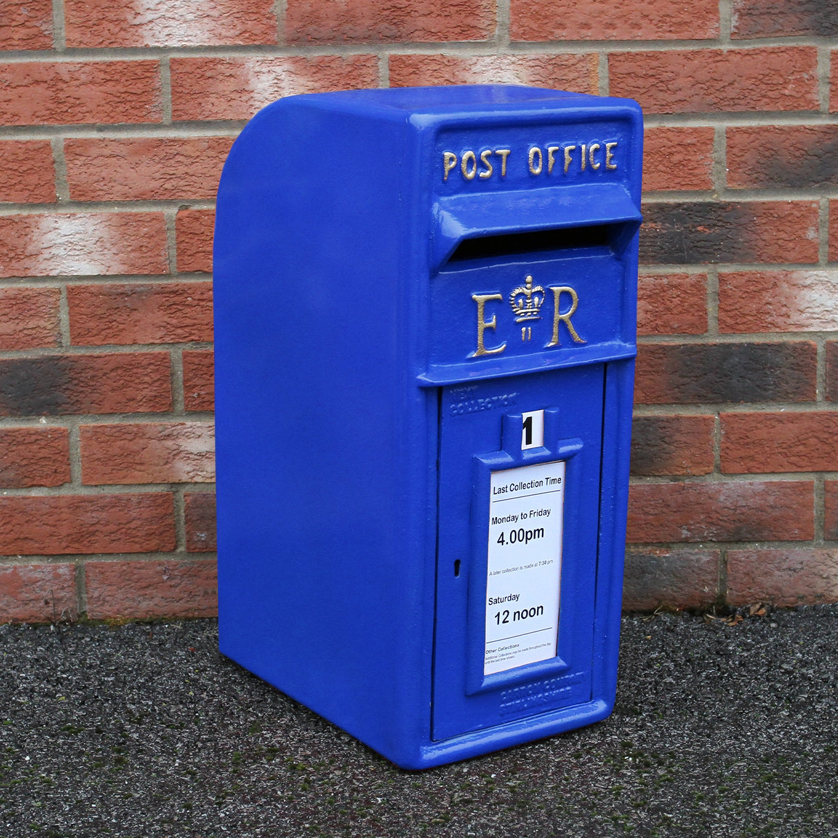 Cassetta postale britannica blu con dettagli dorati, etichetta per orari di raccolta, posizionata su sfondo di mattoni rossi