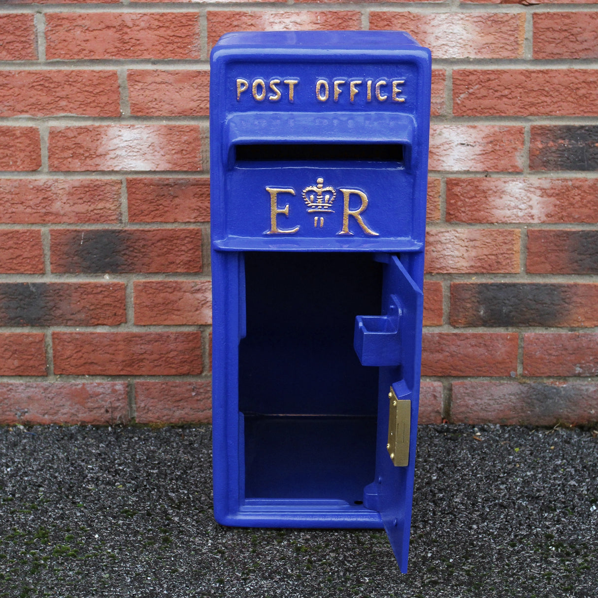 Cassetta postale britannica blu con dettagli dorati, apertura frontale, design vintage e finitura liscia, posizionata su sfondo di mattoni rossi