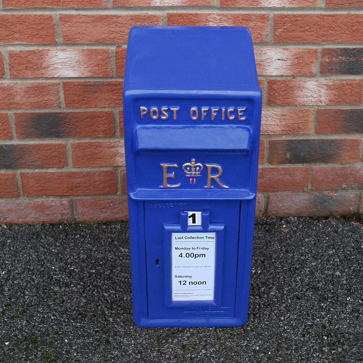Cassetta postale britannica blu con dettagli dorati, logo ER, orari di raccolta e fondo in mattoni rossi e grigi
