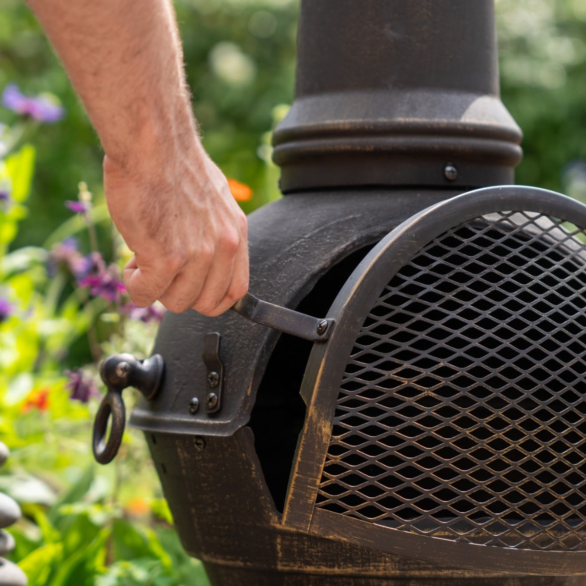 Caminetto bracere da giardino a ciminiera in metallo scuro, con rete protettiva e manico per aprire la porta, circondato da fiori colorati