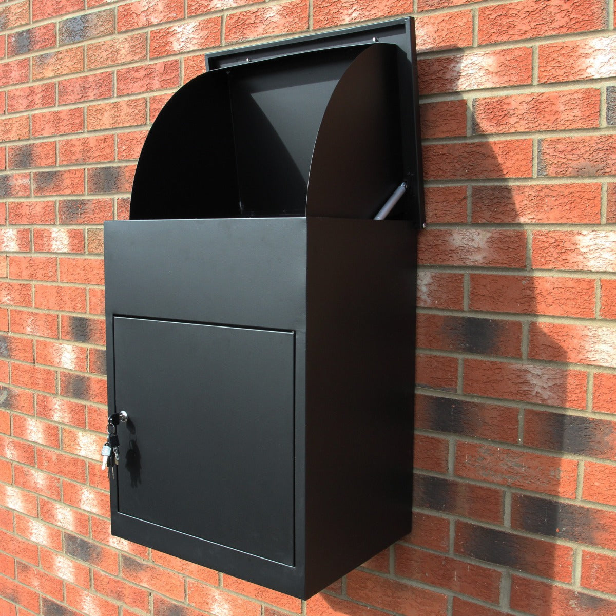Cassetta postale per pacchi nera, con design moderno e finitura opaca, montata su un muro di mattoni rossi, dotata di serratura