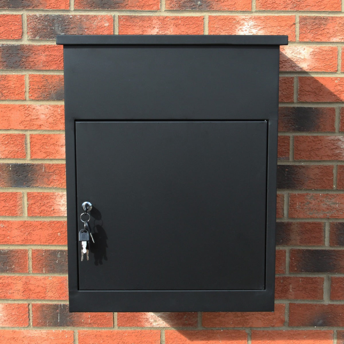 Cassetta postale per pacchi di colore nero, con chiusura a chiave, montata su muro di mattoni rossi, design moderno e funzionale