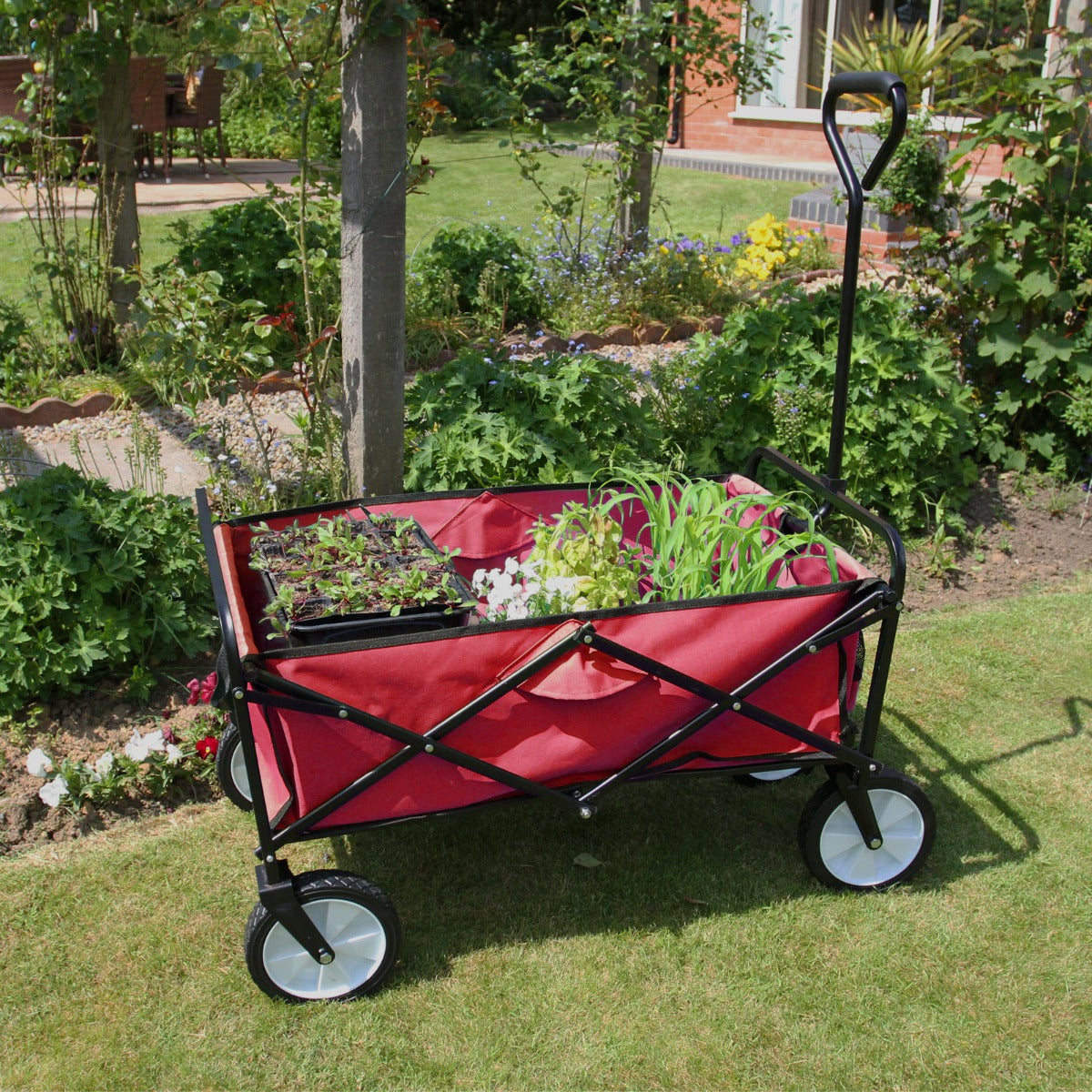 Carrello da giardinaggio pieghevole rosso con ruote bianche, ideale per trasportare piante e attrezzi nel giardino fiorito
