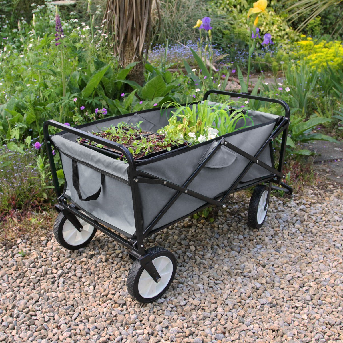 Carrello per giardinaggio pieghevole grigio con ruote bianche, telaio nero e scomparto per piante, posizionato su ghiaia in un giardino fiorito
