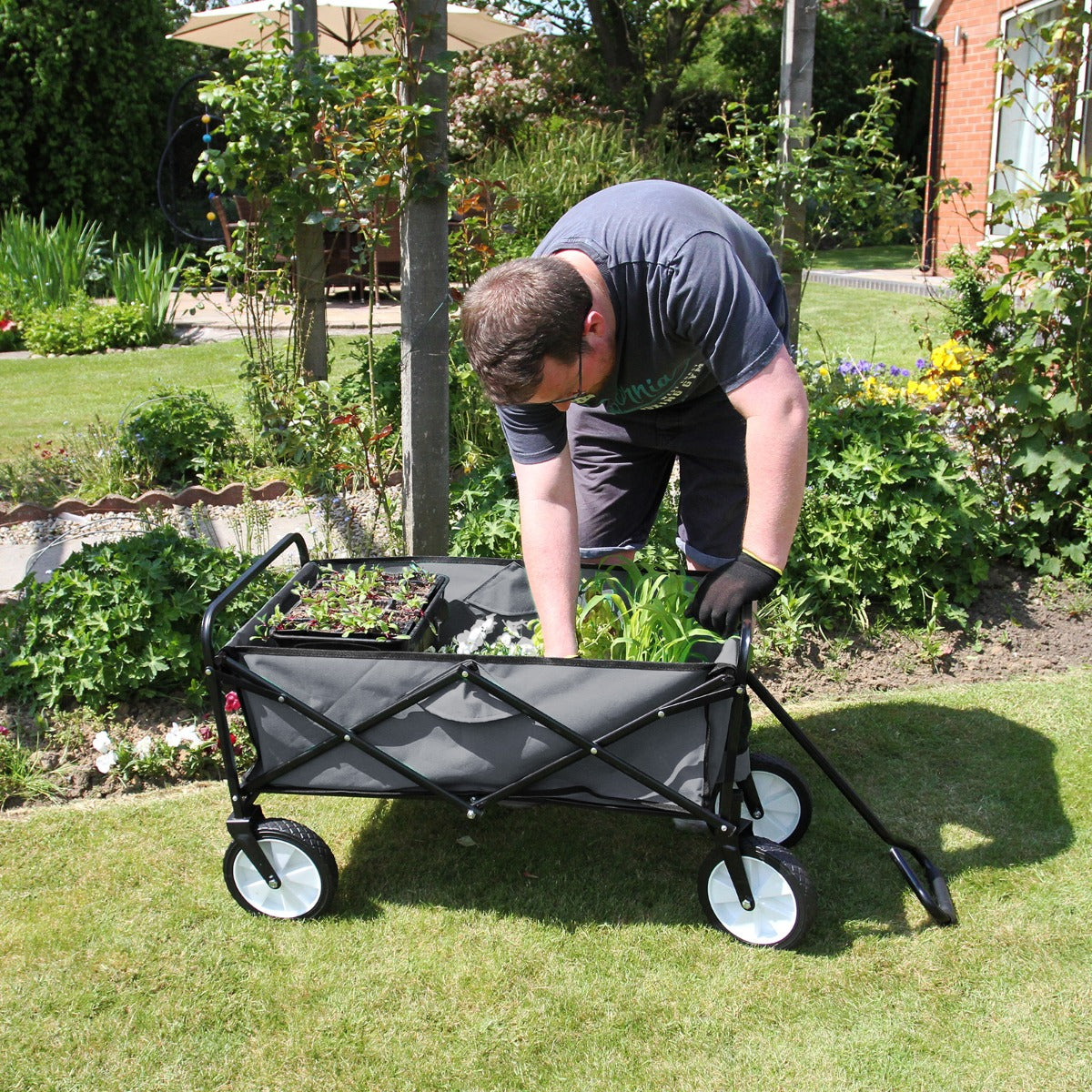 Carrello per giardinaggio pieghevole grigio con ruote bianche, utilizzato per trasportare piante e attrezzi nel giardino verde