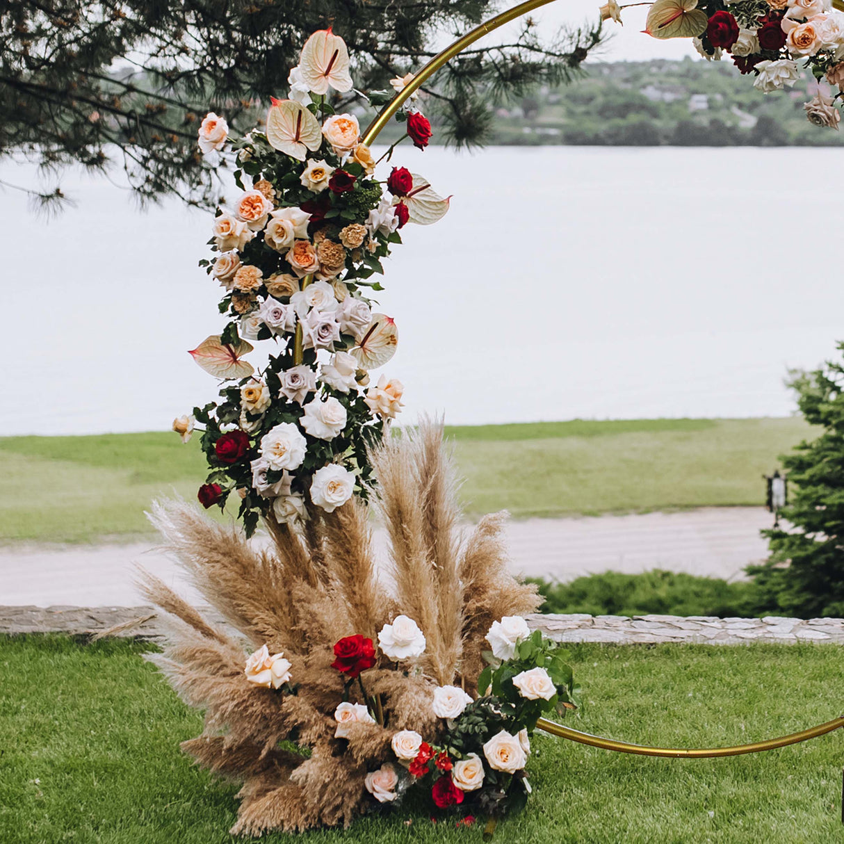 Arco decorativo dorato per matrimoni, adornato con rose bianche, rosse e fiori di pampas, su sfondo verde e acqua serena