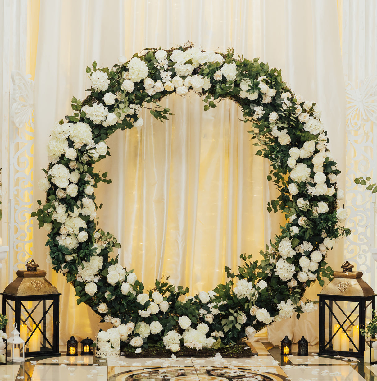 Arco decorativo dorato per matrimoni, adornato con fiori bianchi e verde, circondato da lanterne eleganti su sfondo chiaro